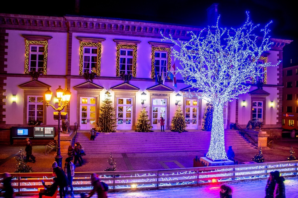 Tree lit up in blue Christmas lights in between a buildings and an ice skating rink with a few skaters