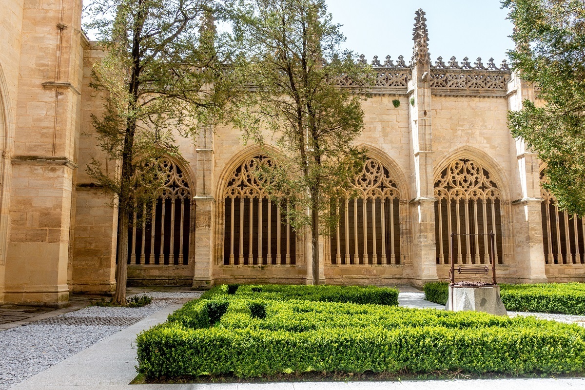 The sunny cloister at the Segovia Cathedral including arched windows and a small hedge in the center