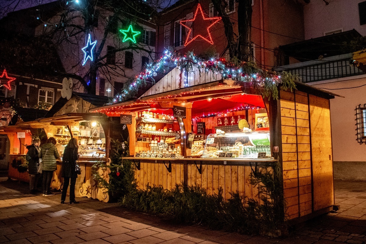 Stand selling cheese that is lit up at night with red and green Christmas lights in Place des Meuniers, part of the Strasbourg Noel market