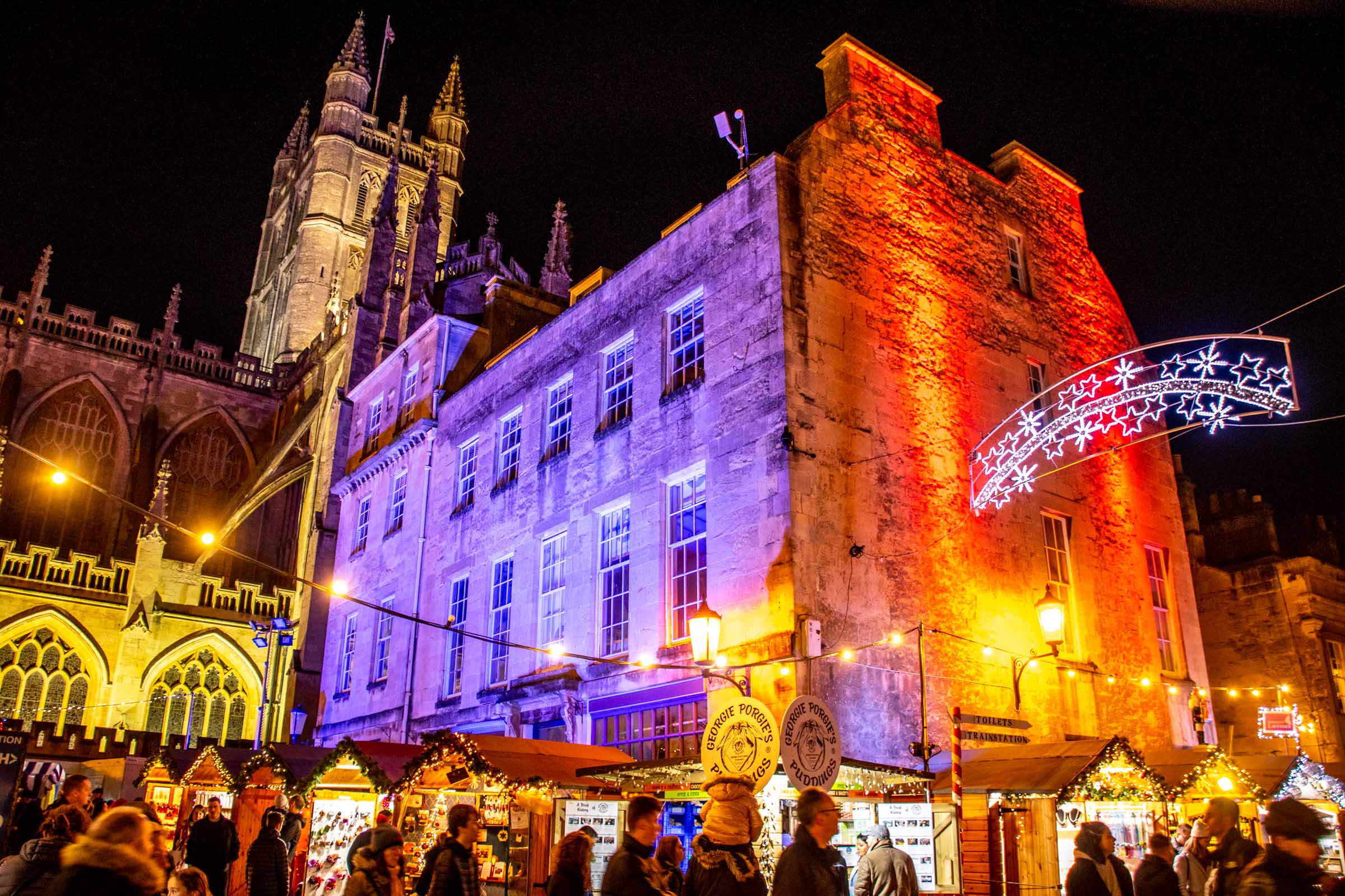 People attending the Bath Christmas market at night in front of Bath Abbey, which is lit up on purple and red lights